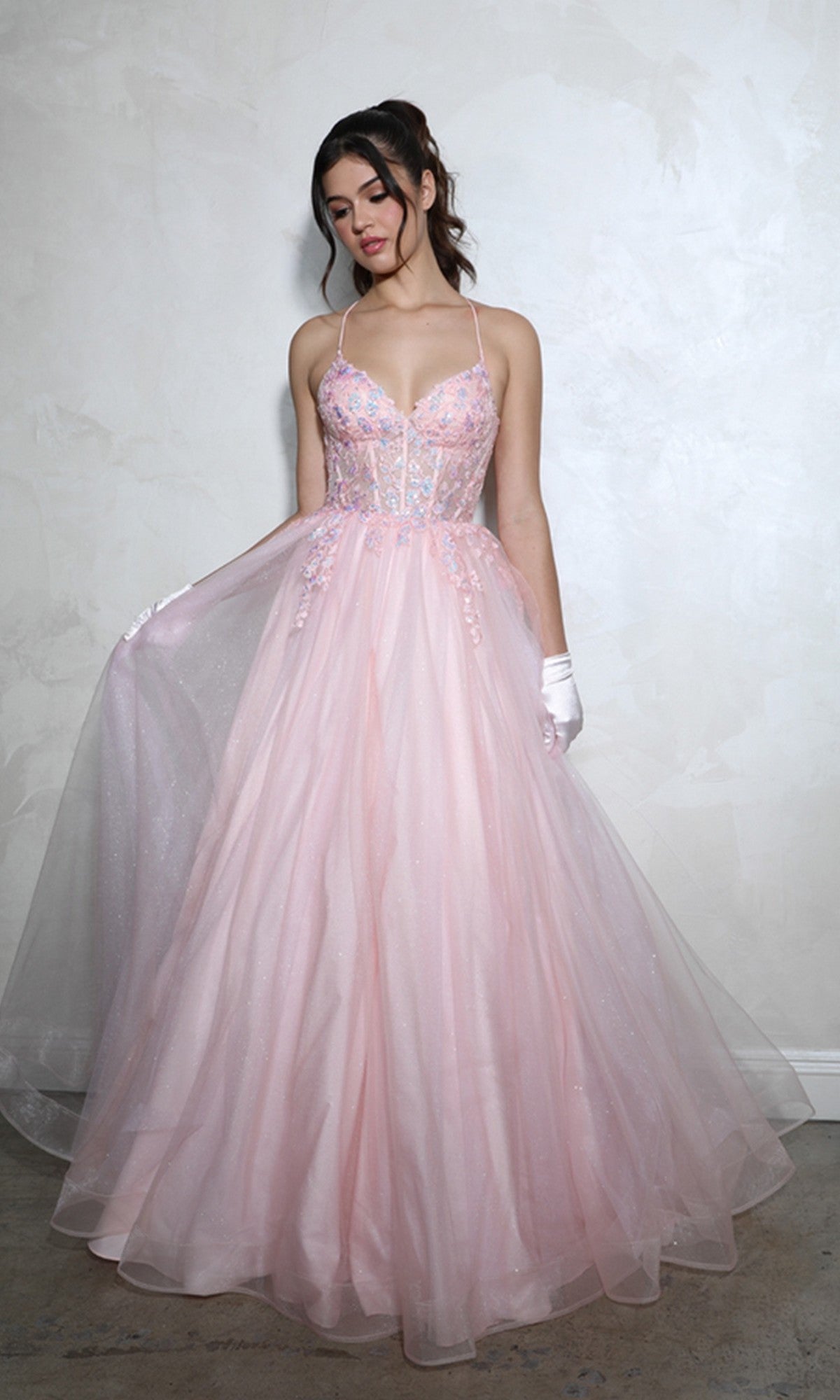 A woman stands against a textured light wall in the pink Lace-Up Corset Long Prom Ball Gown: Eureka 9960, a floor-length, sleeveless dress featuring thin straps and an intricately detailed bodice.